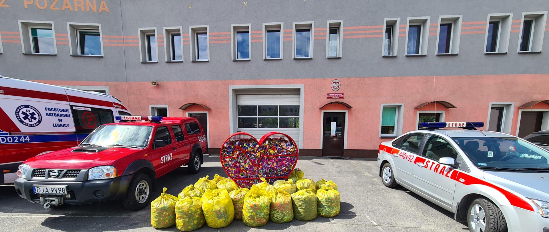 Obraz przedstawia metalowe serce wypełnione plastikowymi nakrętkami. Wokół serca żółte worki wypełnione nakrętkami. Po bokach samochody pożarnicze. W tle budynek komendy.