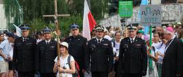 Zdjęcie grupowe uczestników pielgrzymki na tle drzew i fragmentu budynku. Przed pielgrzymami stoi pięciu strażaków w mundurach wyjściowych i czapkach rogatywkach. Przed strażakami stoi kobieta w jasnym ubiorze trzymająca w rękach duży drewniany krzyż. 