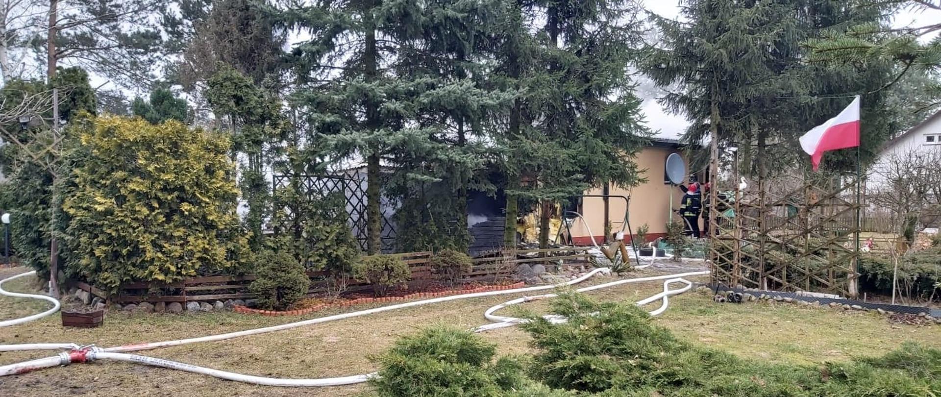 Dom parterowy w trakcie pożaru. Wokół domu rozwinięte linie gaśnicze. Przy jednym oknie stoją strażacy w aparatach ochrony dróg oddechowych.