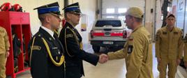 Oficer w ciemnym mundurze wyjściowym ze złotymi sznurami galowymi podaje rękę nowemu funkcjonariuszowi w ramach gratulacji; w tle widoczny jest srebrny samochód strażacki Ford Ranger.