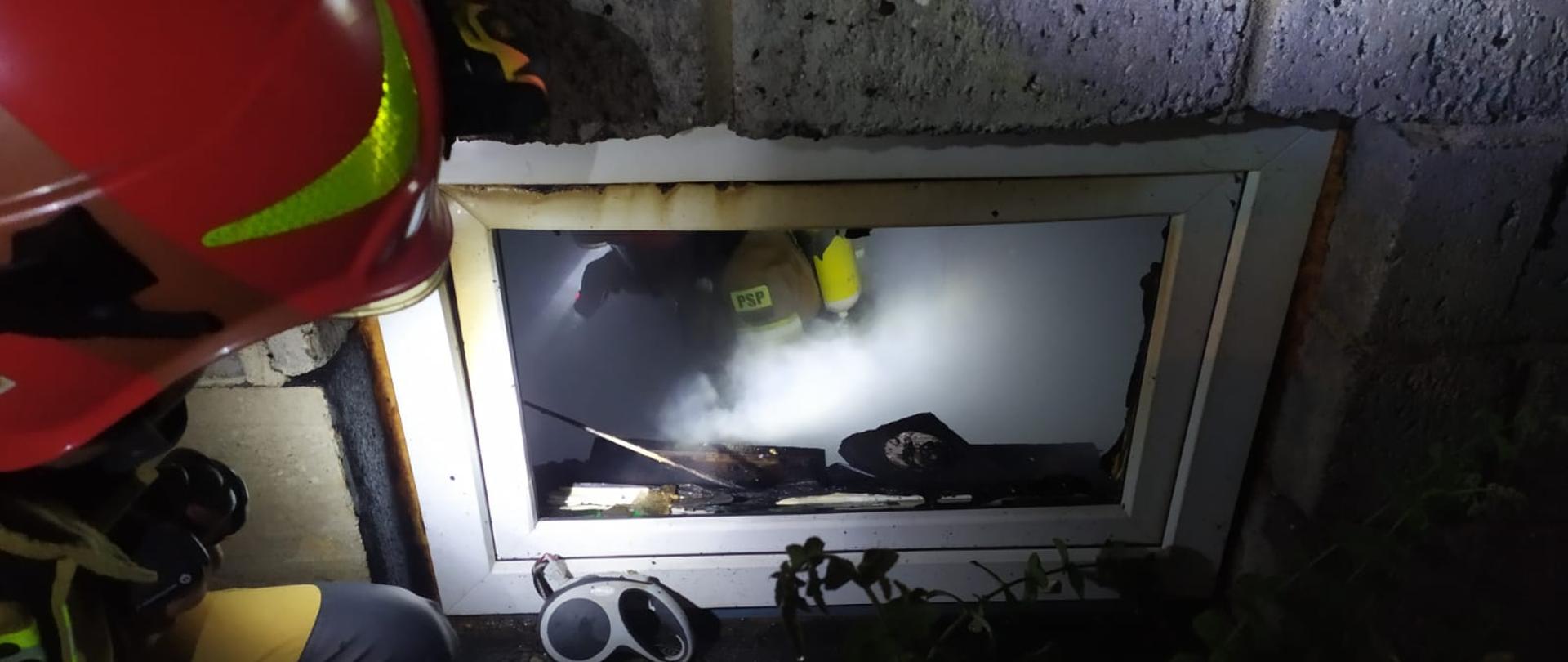 Zdjęcie przedstawia okno z wybitą szybą do pomieszczenia kotłowni. Wewnątrz widoczne zadymienie i strażaka prowadzącego akcję gaśniczą. Drugi strażak klęczy na zewnątrz oświetlając okno ręczną latarką.