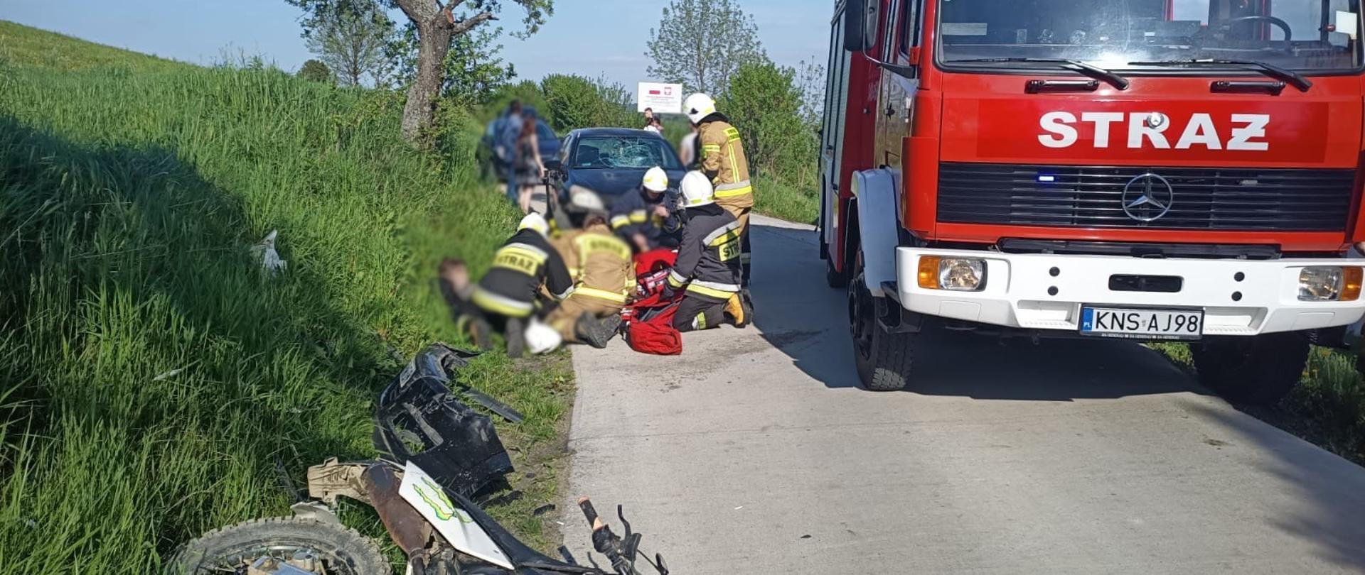 Strażacy udzielają pomocy poszkodowanej osobie leżącej na trawie. Obok stoi samochód pożarniczy