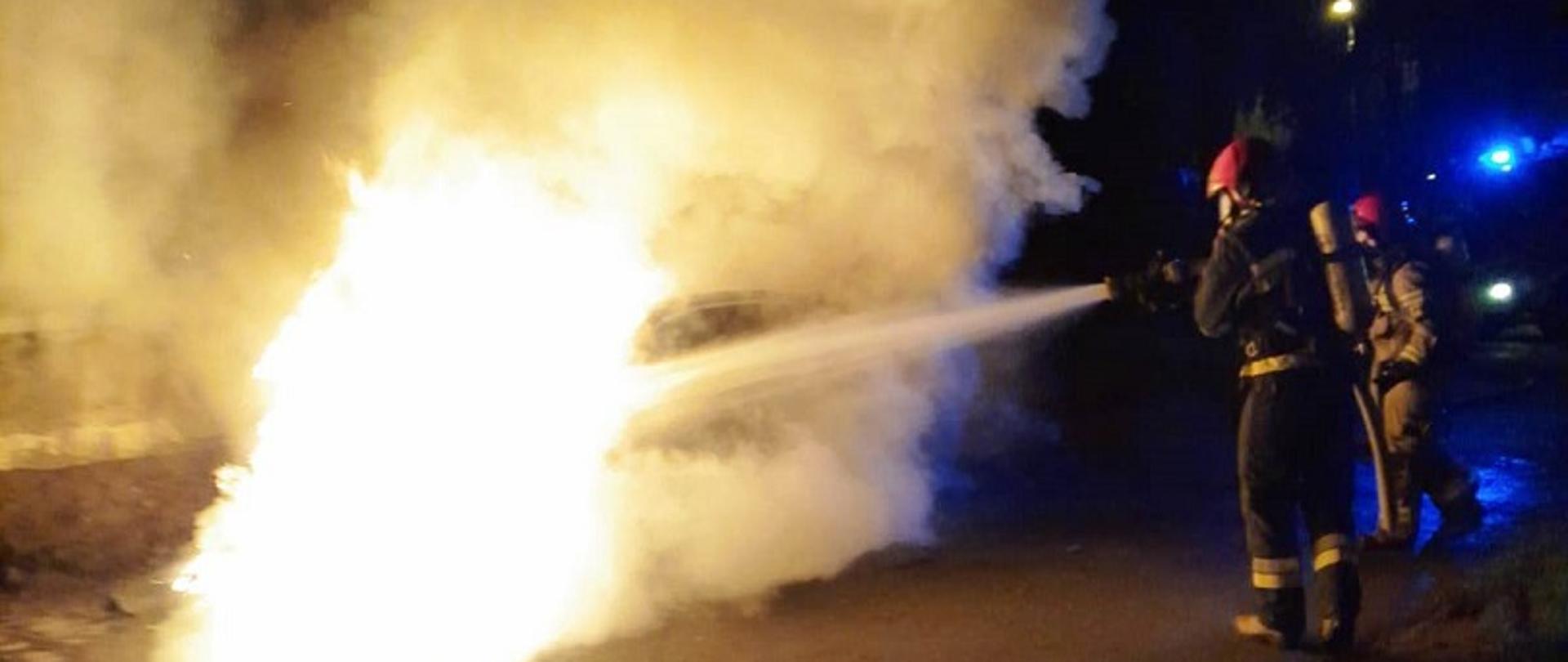 Na zdjęciu widać palący się samochód osobowy marki BMW oraz strażaków ubranych w ubrania bojowe i aparaty ochrony dróg oddechowych podających prąd wody na palący się pojazd. W tle widać sygnały świetlne samochodu pożarniczego.