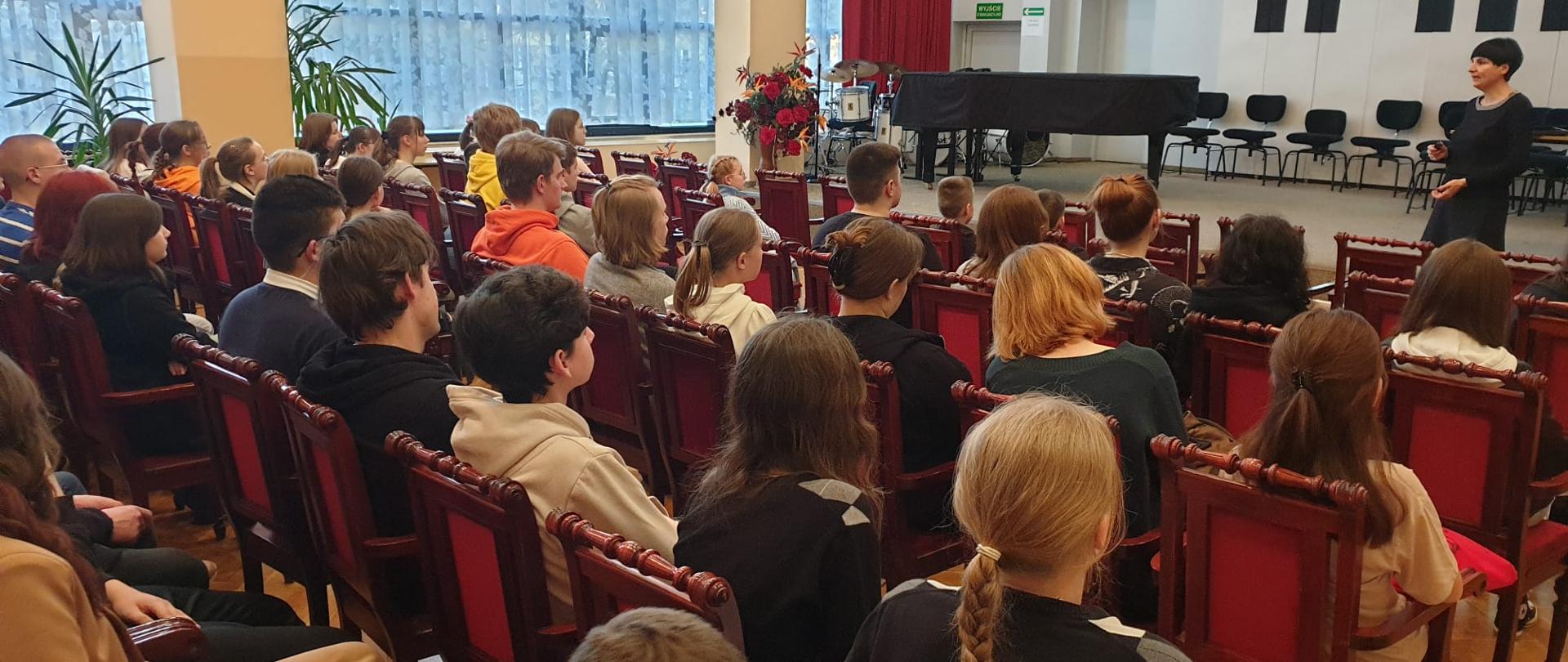 Uczniowie (młodzież) siedzi na krzesłach - ujęcie z boku od tyłu. W tle po prawej stronie widać kobietę prowadząca warsztaty, która mówi stojąc przed sceną z fortepianem.