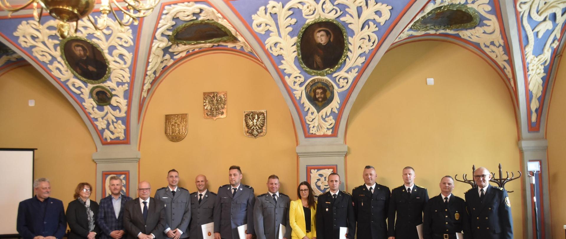 Zdjęcie przedstawia grupę ludzi stojących w rzędzie w zabytkowym pomieszczeniu ze zdobionymi łukami sklepienia. Wśród nich są funkcjonariusze służb mundurowych i osoby cywilne.