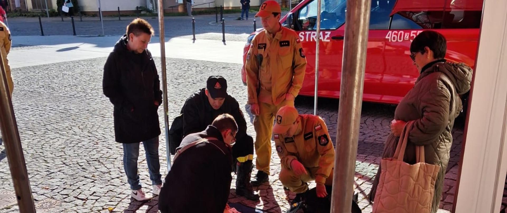 Na zdjęciu widać grupę osób zgromadzonych pod czerwonym namiotem podczas wydarzenia plenerowego. Po lewej stronie stoi mężczyzna w żółtym uniformie z naszywkami i czapką, prawdopodobnie przedstawiciel służb ratowniczych lub organizacji bezpieczeństwa. Rozmawia on z uczestnikami, którzy stoją przy wysokich, okrągłych stołach i wypełniają formularze lub ankiety. Na jednym ze stołów leżą liczne ulotki z ilustracjami i tekstem, a także długopisy. Scena odbywa się na brukowanym dziedzińcu przy ceglanej ścianie budynku.