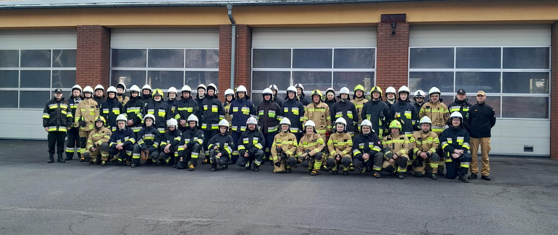 Na zdjęciu znajduje się 41 absolwentów szkolenia podstawowego strażaka ratownika OSP i trzech członków komisji egzaminacyjnej. Kursanci ubrani w ubrania specjalne. Zdjęcie zrobione na tle bram garażowych JRG w Oleśnie.
