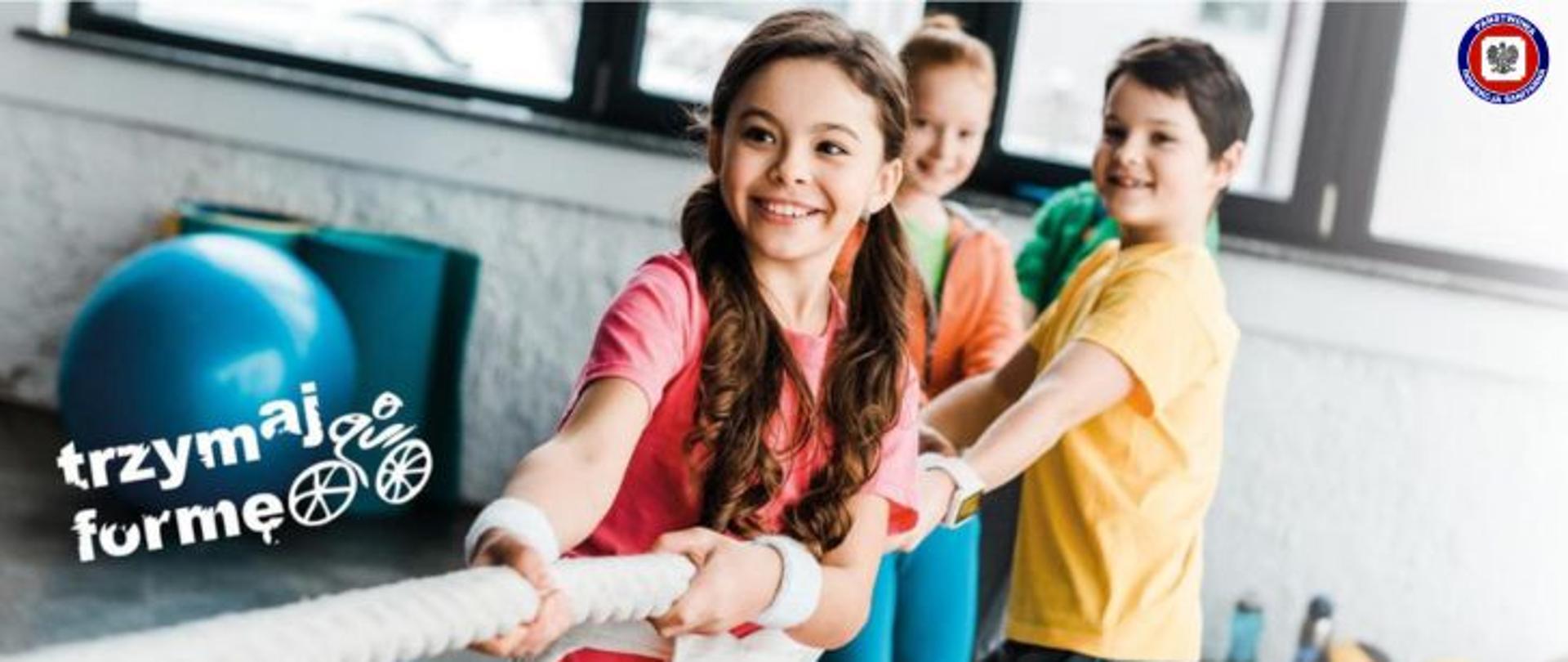 Uśmiechnięte dzieci na sali gimnastycznej ciągną linę, po ich lewej napis trzymaj formę. W prawym górnym rogu logo Państwowej Inspekcji Sanitarnej. 