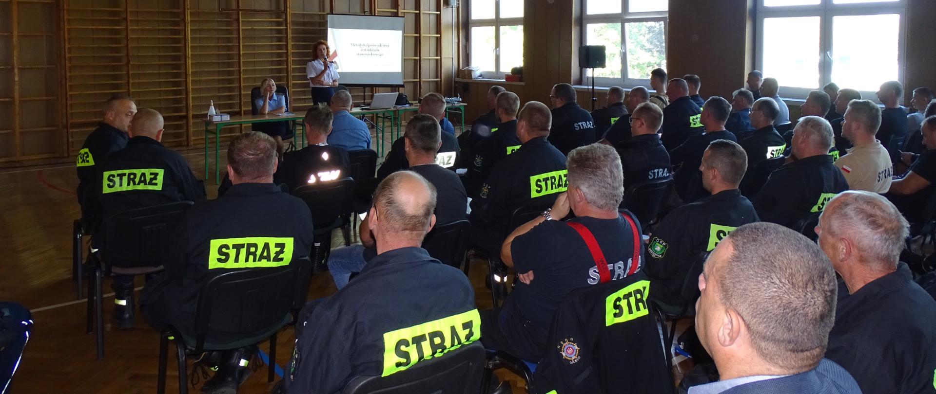 Strażacy OSP siedzą odwróćeni tyłem na krzesłach w sali gimnastycznej, W tle na ekranie wyswietla się prezezntacja, obok za stołem stoi z mikrofonem funkcjonariuszka PSP w białej koszuli, druga siedzi obok za stołem. 