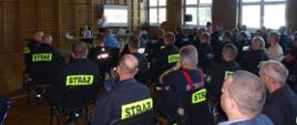 Strażacy OSP siedzą odwróćeni tyłem na krzesłach w sali gimnastycznej, W tle na ekranie wyswietla się prezezntacja, obok za stołem stoi z mikrofonem funkcjonariuszka PSP w białej koszuli, druga siedzi obok za stołem. 