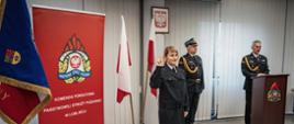 Zdjęcie zrobione w pomieszczeniu strażnicy na tle godła Rzeczpospolitej Polskiej, Sztandaru Komendy Powiatowej PSP w Lublińcu, dwóch Flag Rzeczpospolitej Polskiej oraz po lewej stronie Sztandaru Komendy Powiatowej PSP w Lublińcu, banneru Komendy Powiatowej PSP w Lublińcu. Na pierwszym planie nowo przyjęty strażak stojący na baczność i ubrany w mundur służbowy składa ślubowanie podnosząc prawą rękę, po prawej stronie Komendant w ubraniu galowym stojący za mównicą
