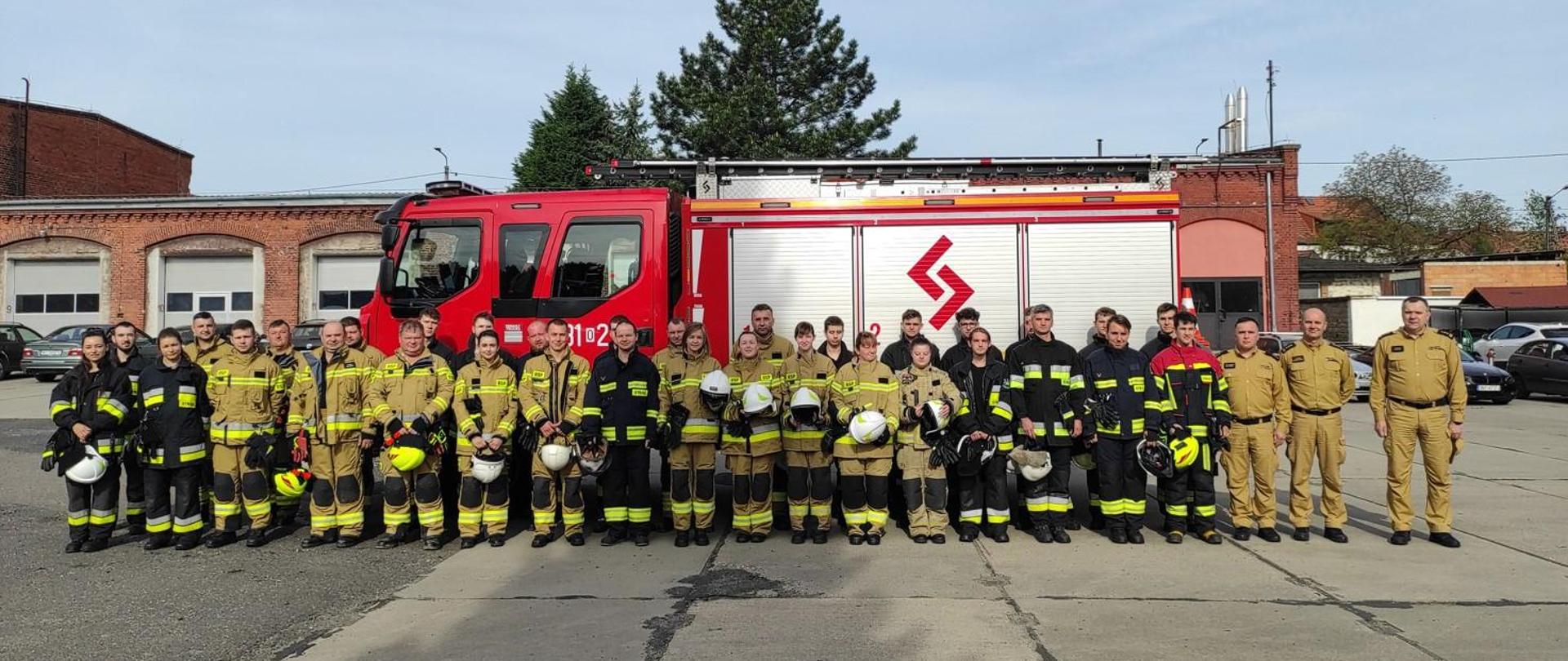 Zdjęcie przedstawia grupę strażaków w mundurach bojowych na placu wewnętrznym, w tle samochód pożarniczy oraz budynek komendy powiatowej PSP.