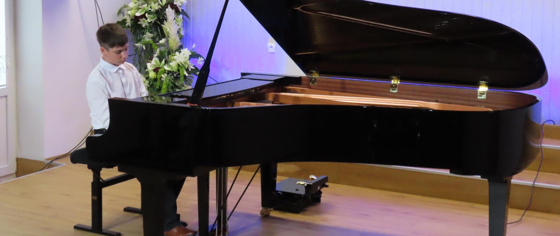 na zdjęciu, w auli PSM widnieje uczeń, który gra na fortepianie podczas konkursu na utwór dowolny. Kolorystyka zdjęcia jest biało-szaro-niebiesko-zielono-żółto-brązowo- czerwono-czarna.