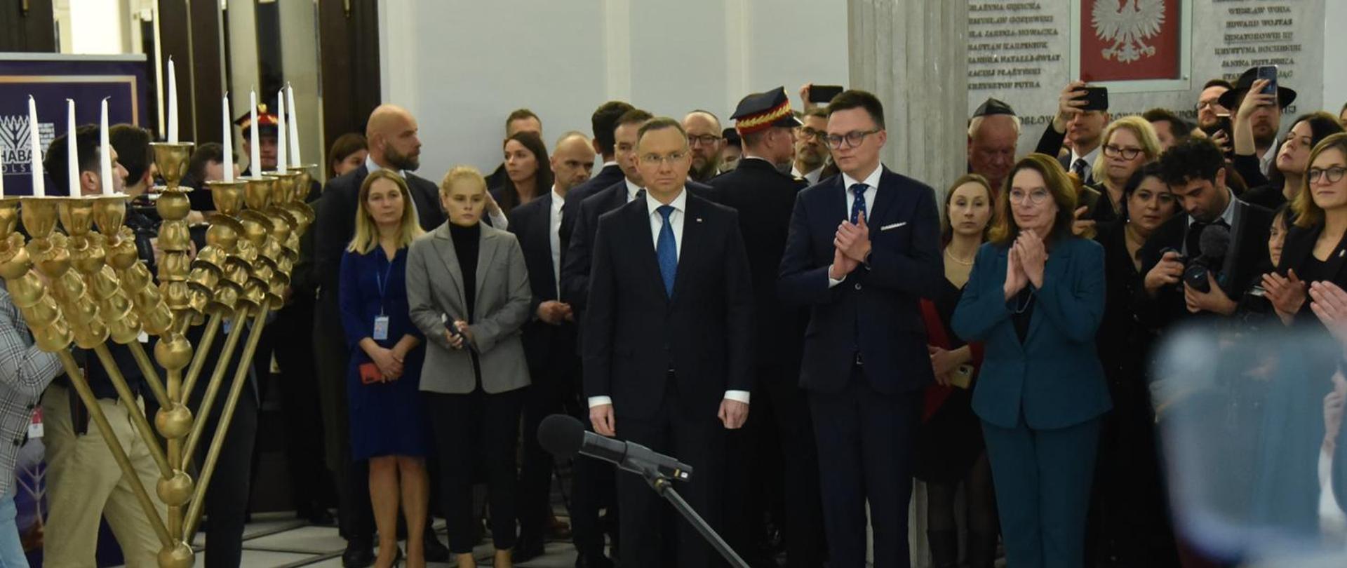 W białej sali stoi dużo ludzi, pośrodku marszałek Hołownia i prezydent Duda, przed nimi złoty dziewięcioramienny świecznik.