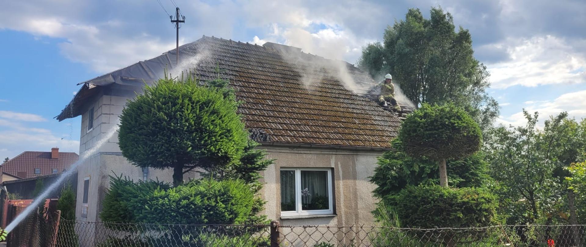 Budynek murowany jednorodzinny, widok za płotu z siatki ogrodzeniowej. Widoczne unoszący się dym nad dachem, dach częściowo rozebrany z dachówki, na dachu na opartej drabinie pracującą strażacy. Widoczny podawany prąd wody na dach.