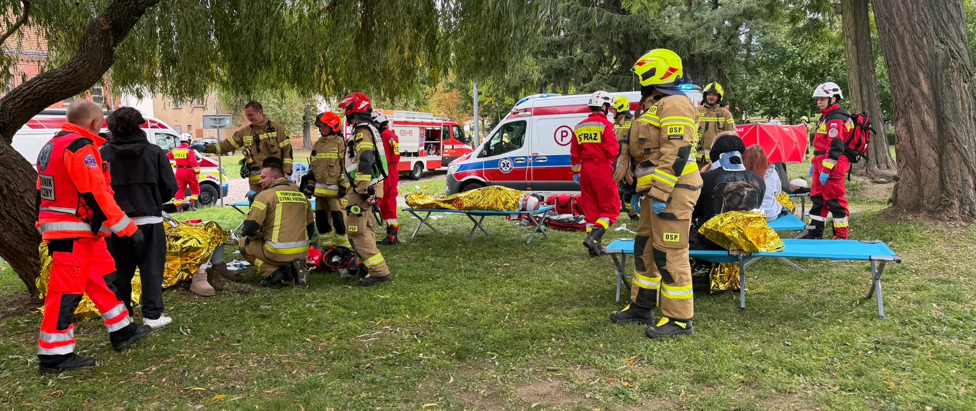Strażacy i ratownicy medyczni udzielają pomocy osobom poszkodowanym w wyznaczonej strefie medycznej. Część osób znajduje się na noszach i jest okryta foliami termicznymi. W tle stoją pojazdy ratownictwa medycznego i straży pożarnej.