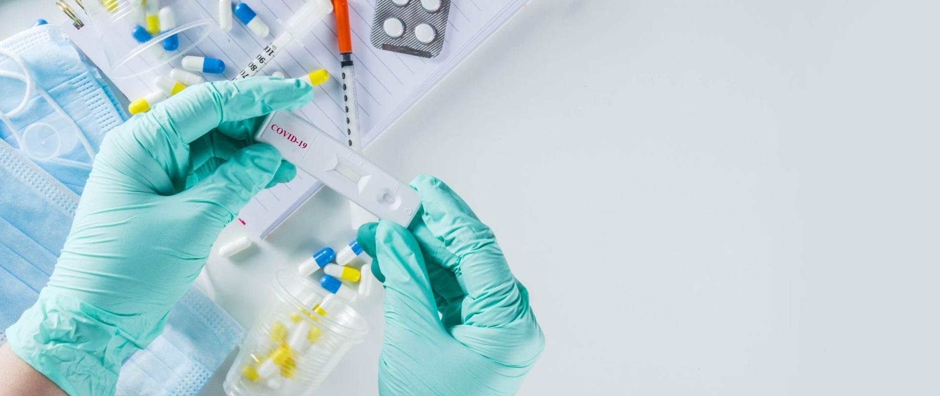 Test na COVID-19 trzymany przez dwie ręce w gumowych rękawiczkach. Pod dłońmi zeszyt w linie, lekarstwa, maseczka