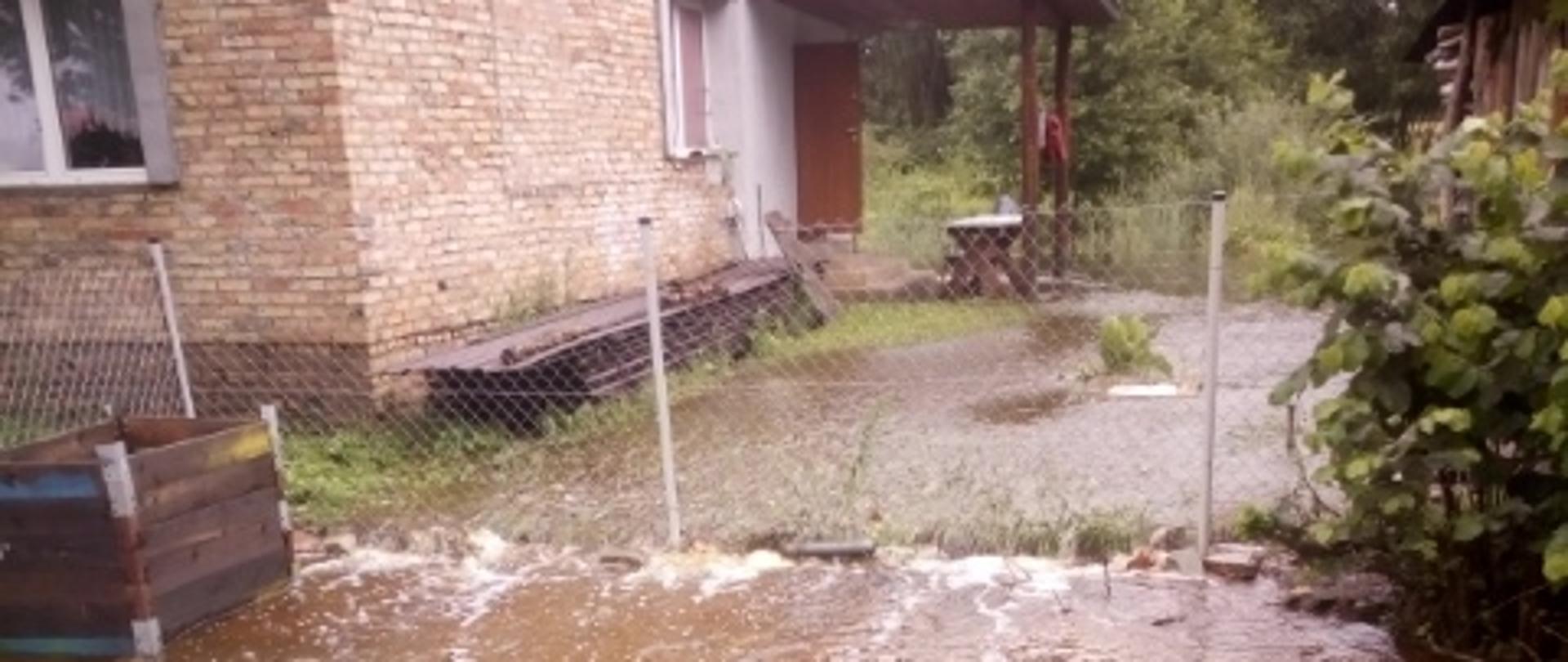 Na zdjęciu zalana posesja w oddali dom z czerwonej cegły i ogrodzenie z siatki metalowej.