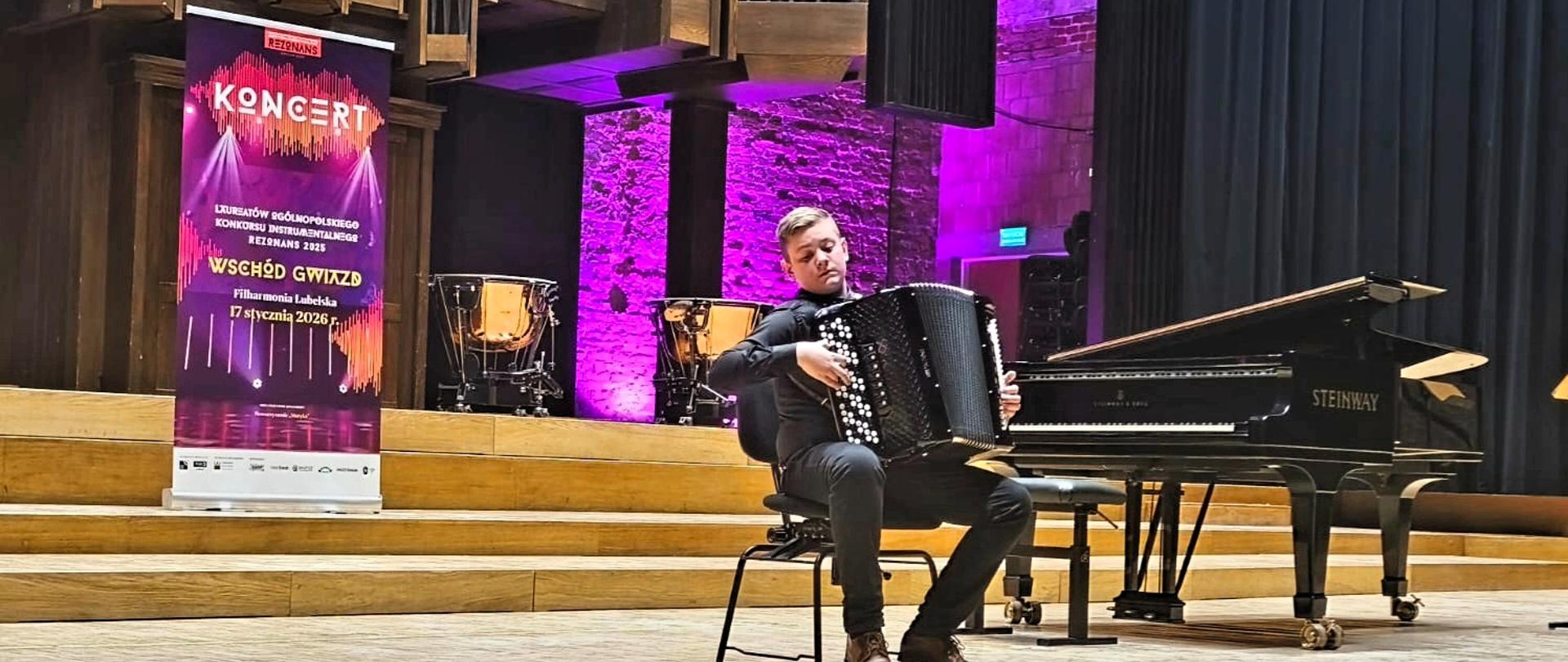 Szeroki plan na scenę Filharmonii Lubelskiej. Na środku siedzi młody muzyk grający na akordeonie. Obok niego stoi czarny fortepian marki Steinway oraz wysoki roll-up koncertowy. W tle monumentalne organy i kotły symfoniczne, podświetlone na fioletowo.