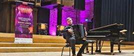 Szeroki plan na scenę Filharmonii Lubelskiej. Na środku siedzi młody muzyk grający na akordeonie. Obok niego stoi czarny fortepian marki Steinway oraz wysoki roll-up koncertowy. W tle monumentalne organy i kotły symfoniczne, podświetlone na fioletowo.