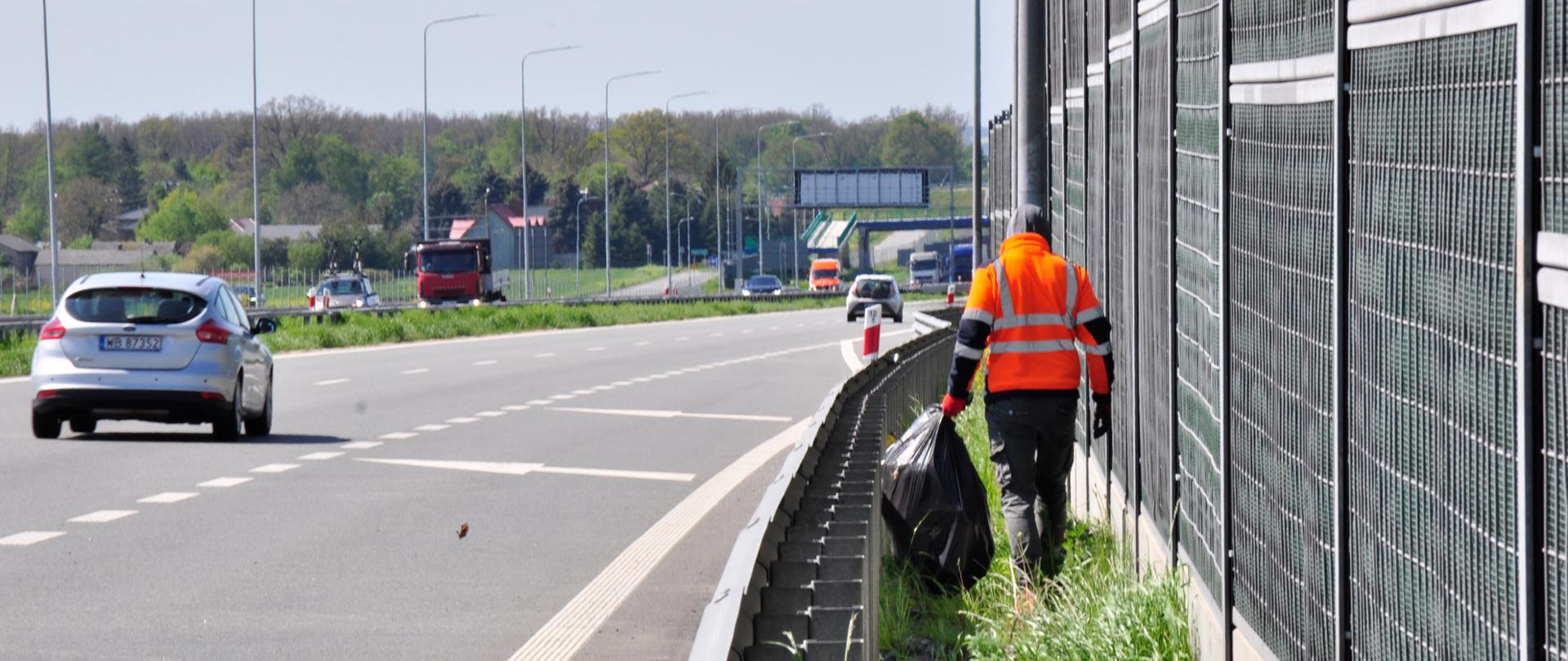 Zdjęcie przedstawia osobę ubraną w pomarańczową kamizelkę odblaskową, która porusza się wzdłuż pobocza autostrady, zbierając śmieci. Osoba ta trzyma duży czarny worek na śmieci i idzie obok metalowego ogrodzenia dźwiękochłonnego. Na drodze w tle widoczne są pojazdy, w tym srebrny samochód osobowy oraz czerwona ciężarówka. Po lewej stronie zdjęcia znajduje się pas zieleni oddzielający jezdnię od pobocza. W tle widoczne są drzewa oraz budynki, a dzień jest pogodny, z lekkim zachmurzeniem.