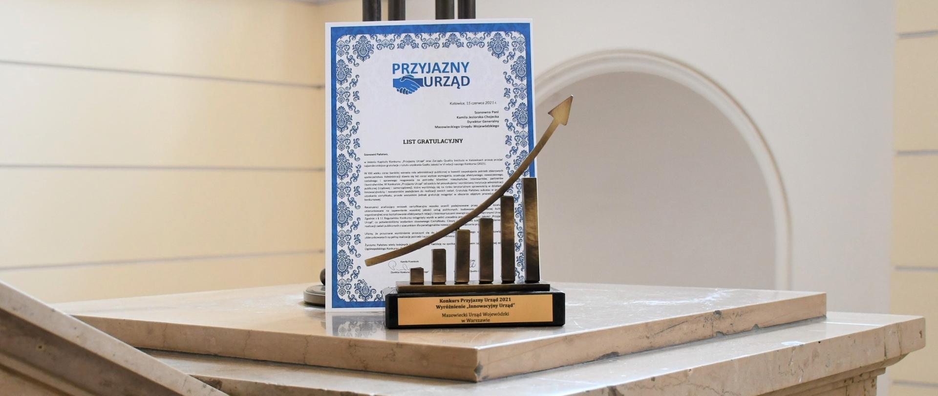 Na zdjęciu znajduje się złota statuetka (wzrastające słupki ze strzałą skierowaną do góry) oraz biało niebieski list gratulacyjny