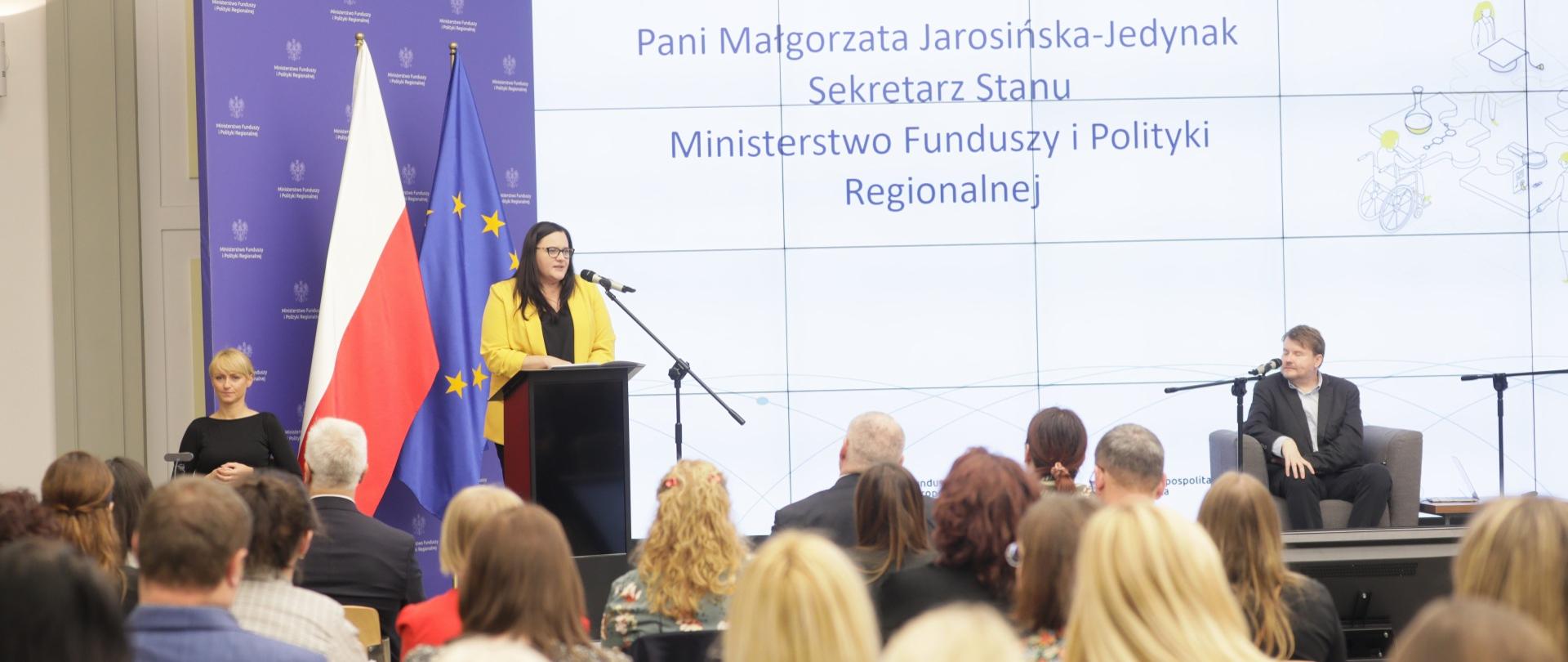 Wiceminister Małgorzata Jarosińska-Jedynak w mównicy z mikrofonem. Za nią flagi PL i UE i ekran. Przed publiczność siedząca na krzesłach.
