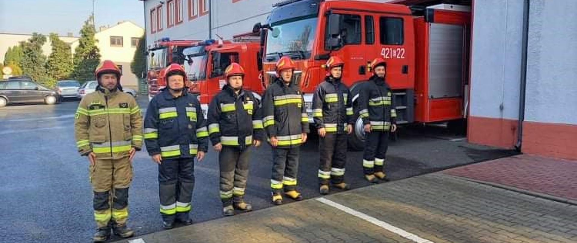 Przed strażnicą w szyku na baczność stoją strażacy w ubraniach specjalnych i czerwonych hełmach. Za nimi widać trzy wozy strażackie, które wyjechały z garażu strażnicy.