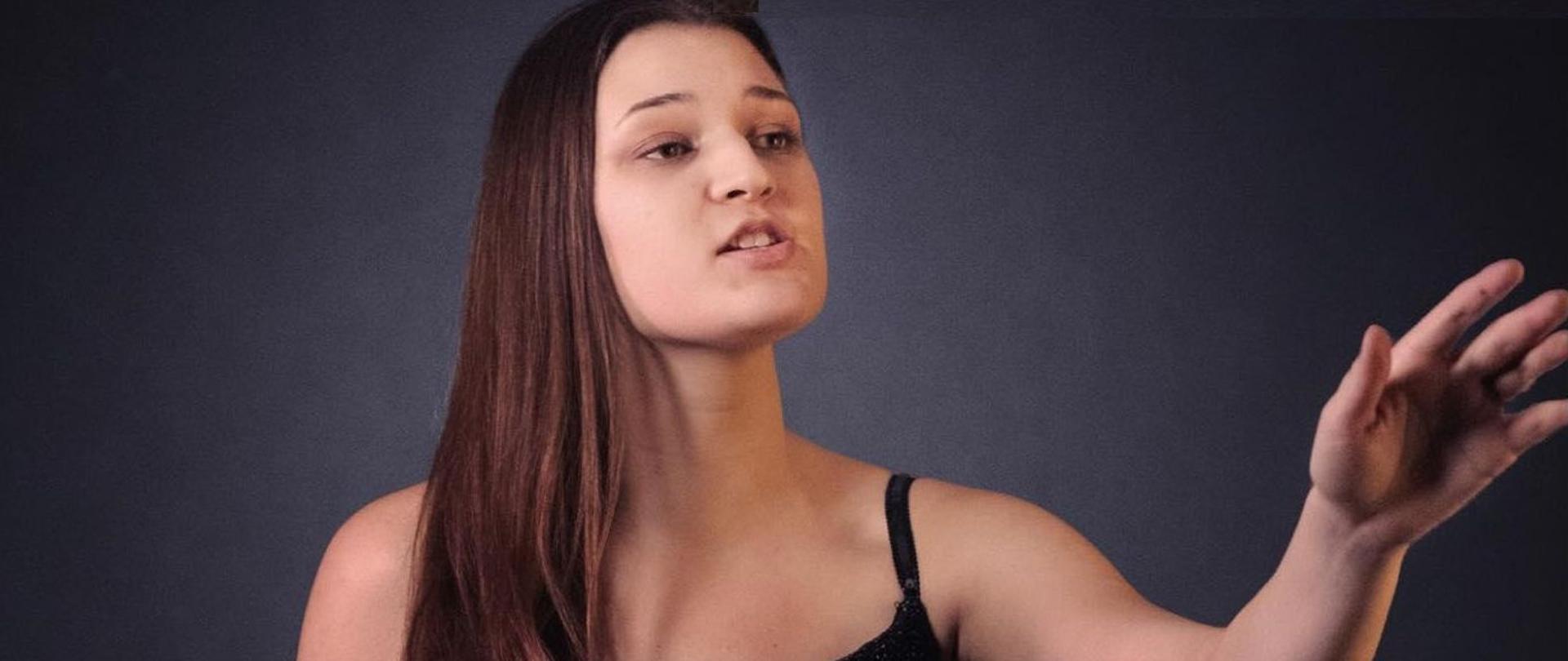 Zdjęcie kolorowe. Dziewczyna w stroju wieczorowym. Śpiewa. Stoi z wyciągniętą w przód lewą ręką.