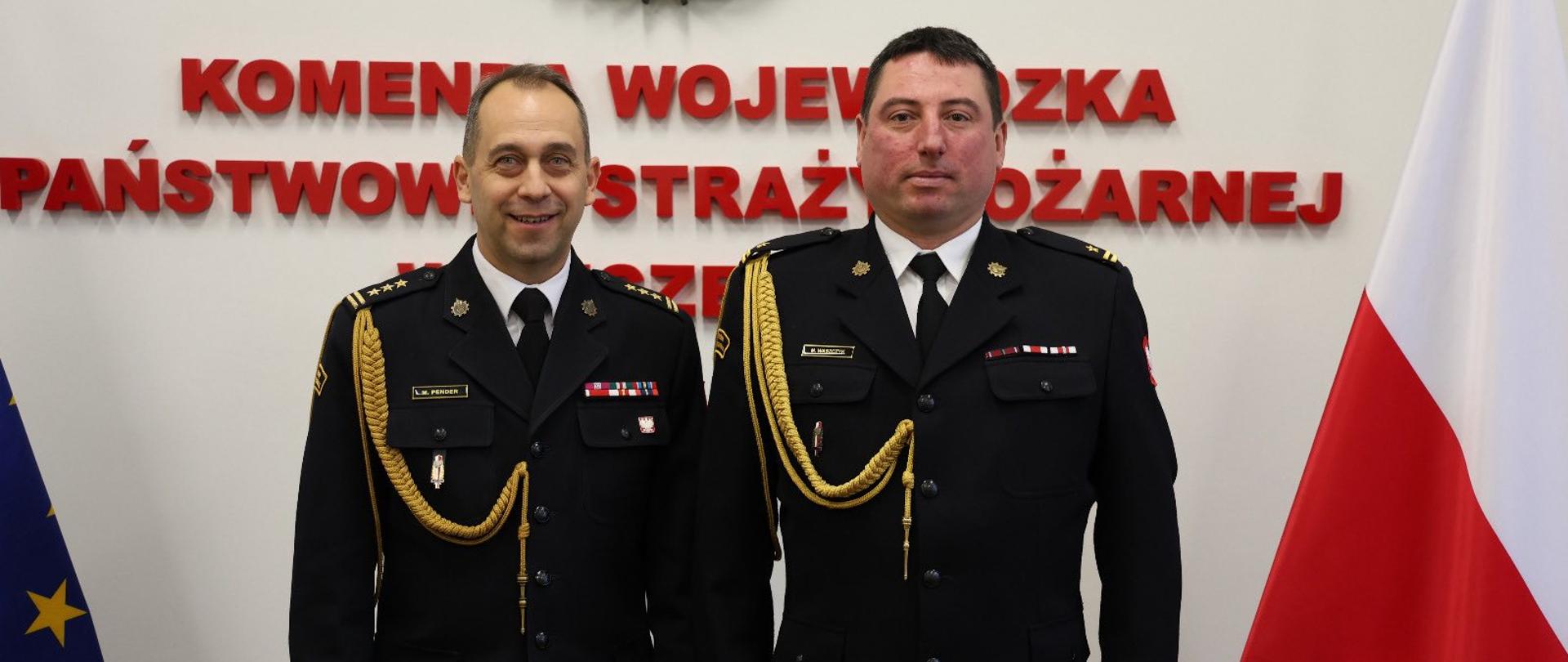 Na zdjęciu stoją obok siebie dwaj mężczyźni w galowych mundurach straży pożarnej. Pozują do zdjęcia na tle ściany z napisem w języku polskim: „Komenda Wojewódzka Państwowej Straży Pożarnej”. Nad napisem znajduje się emblemat straży pożarnej. Obaj mężczyźni mają ciemne, granatowe mundury z pagonami oraz ozdobnymi, złotymi sznurami. Po lewej stronie stoi uśmiechnięty mężczyzna o krótkich, jasnych włosach. Po prawej stronie stoi drugi mężczyzna, z krótkimi, ciemnymi włosami, z poważnym wyrazem twarzy. Po bokach zdjęcia widoczne są dwie flagi – po lewej flaga Unii Europejskiej, po prawej flaga Polski.