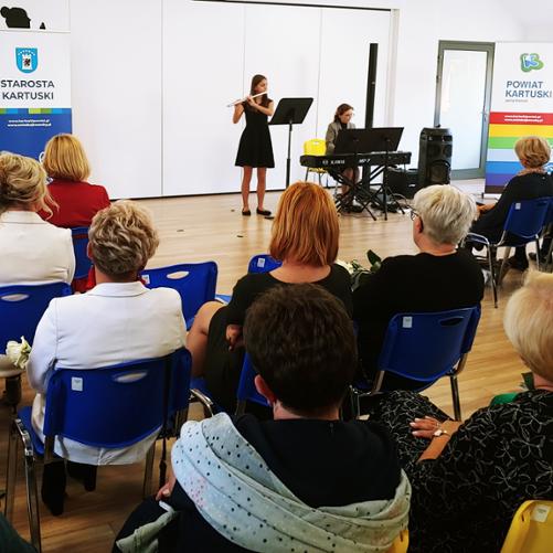 zdjęcie przedstawia uczennicę grającą na flecie przy akompaniamencie pianistycznym nauczycielki, widoczni są siedzący słuchacze