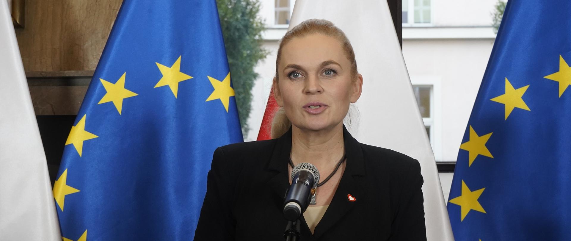 Minister Nowacka stoi na tle rzędu flag Polski i UE i mówi do mikrofonu na stojaku.