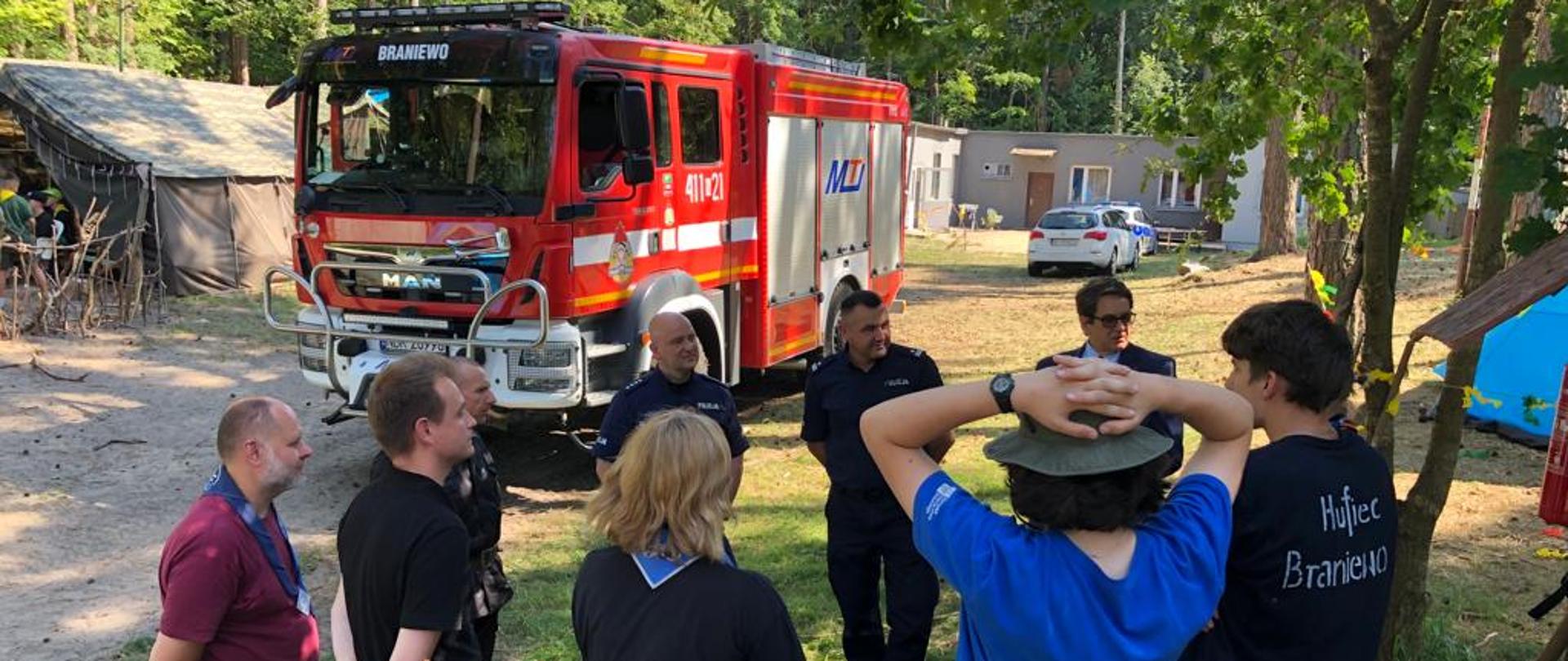 na polanie w lesie stoi duży czerwony samochód strażacki. przed nim grupa mundurowych i harcerzy. Słonecznie.