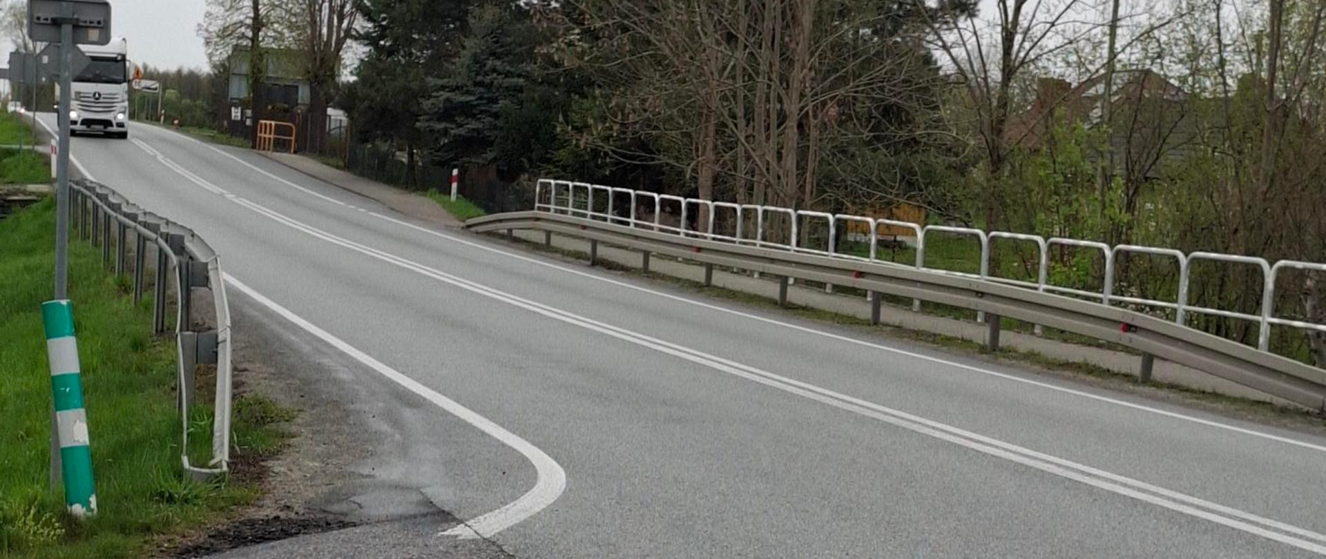 DK73 zjazd w Żelazówce na Odporyszów, widoczny ruch pojazdów ciężkich, brak pobocza i chodników oraz gęstą zabudowę