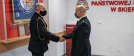 Zdjęcie przedstawia dwóch strażaków komendy miejskiej w mundurach wyjściowych koloru czarnego na tle czerwonego napisu Komenda Miejska Państwowej Straży Pożarnej w Skierniewicach. Jeden ze strażaków gratuluje drugiemu poprzez podanie dłoni.