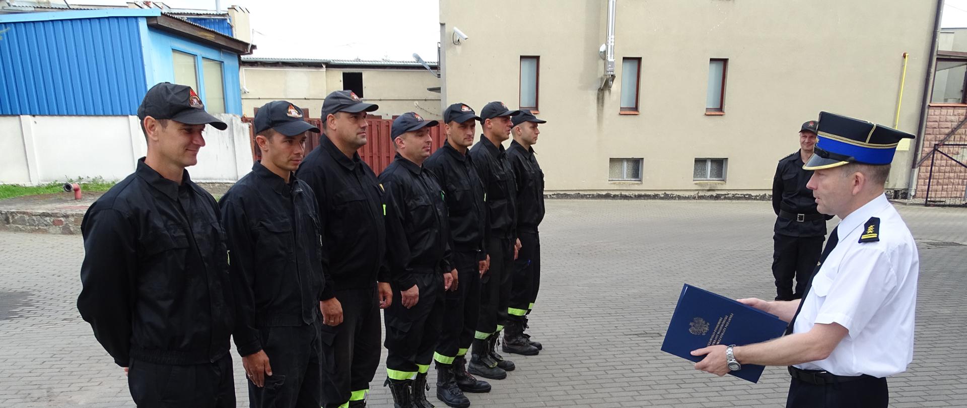 Zdjęcie wykonano w ciągu dnia na placu Komendy Powiatowej Państwowej Straży Pożarnej w Sępólnie Krajeńskim. Strażacy Państwowej Straży Pożarnej ubrani w mundury koszarowe stoją w szeregu przed Komendantem Powiatowym Państwowej Straży Pożarnej ubranym w mundur wyjściowy trzymającym w ręku decyzję Komendanta Wojewódzkiego. 