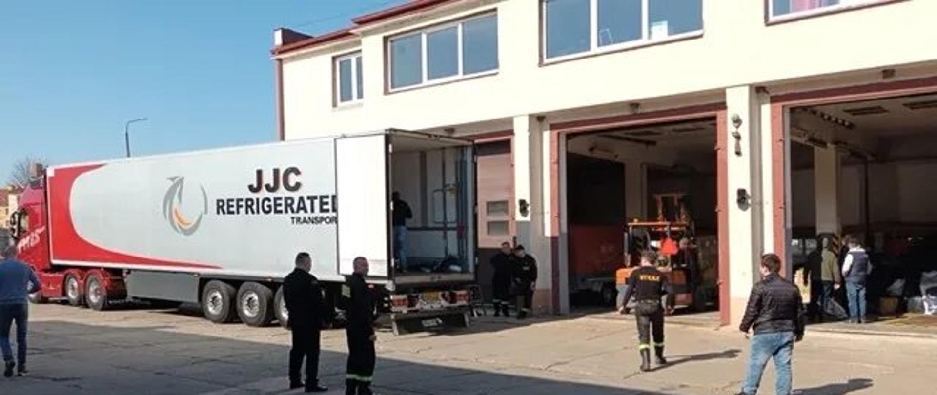 Fotografia przedstawia samochód ciężarowy zaparkowany na terenie Komendy Powiatowej Straży Pożarnej w Malborku, z którego strażacy rozładowują dary dla uchodźców z Ukrainy do jednego z garaży.