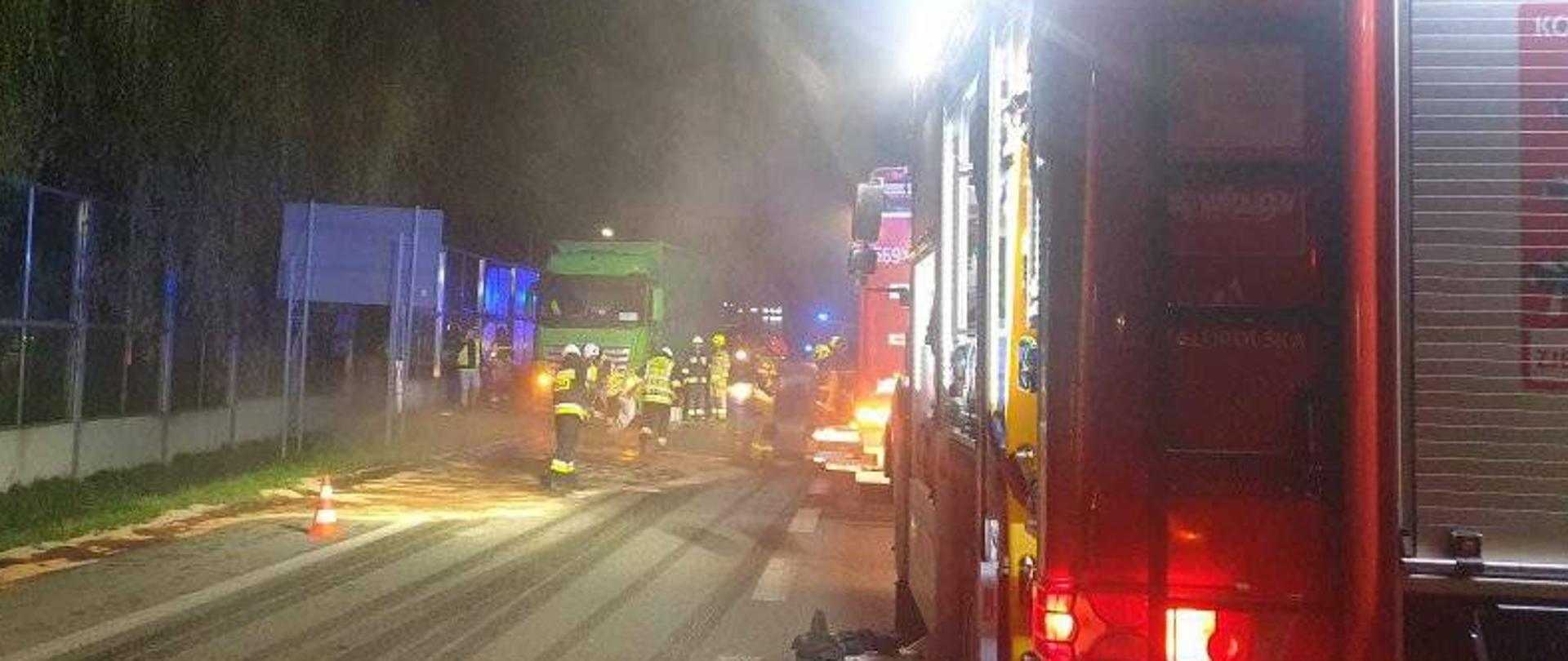 Na zdjęciu widać część samochody pożarniczego. W dalszej części na jezdni samochód ciężarowy oraz strażaków