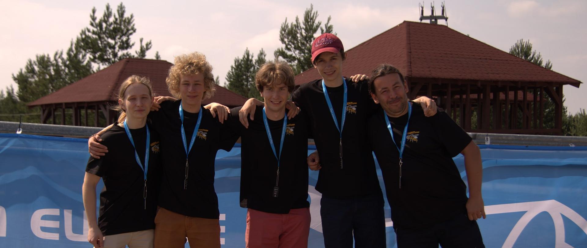 Grupa chłopców stoi na plaży i trzyma się za ramiona. Z nimi niebieski parawan. Są ubrani w czarne koszulki. Na szyi mają zawieszone błękitne smycze. W tle drzewa i altany. 