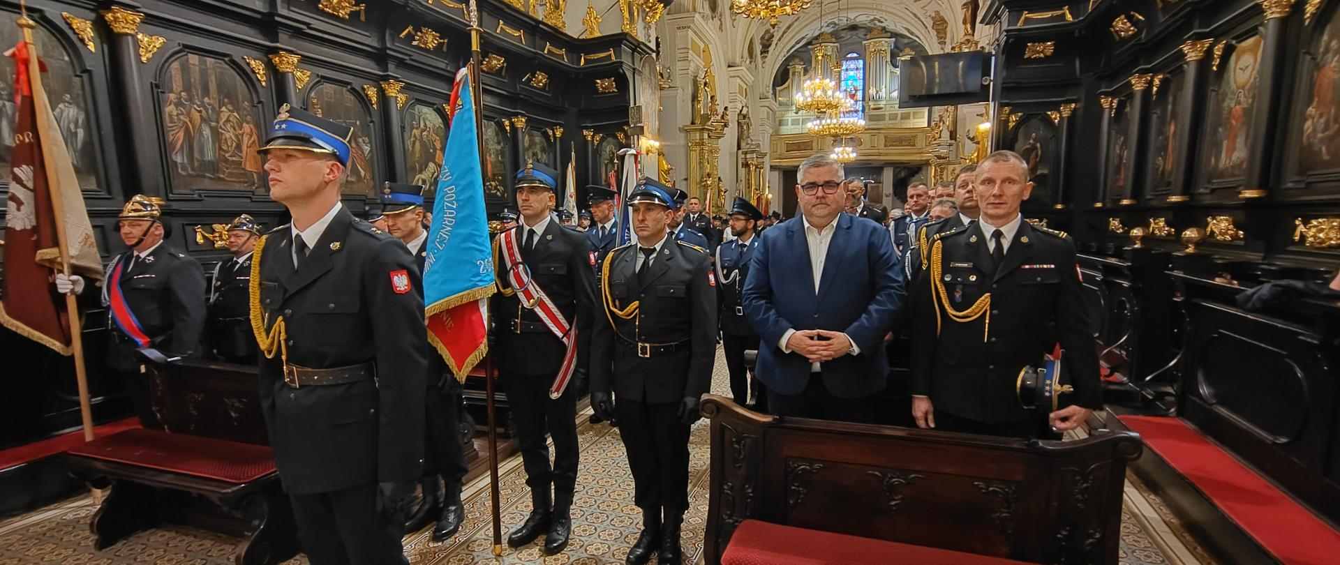 Uroczysta msza w bogato zdobionym, barokowym wnętrzu kościoła. Przed drewnianymi ławami stoją na baczność strażacy w mundurach galowych, poczet sztandarowy z niebieskim sztandarem oraz mężczyzna w garniturze.