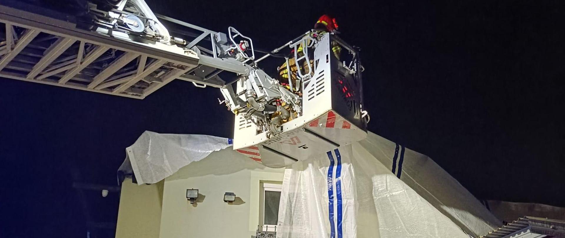 Strażak na drabinie mechanicznej przykrywa uszkodzony dach folią