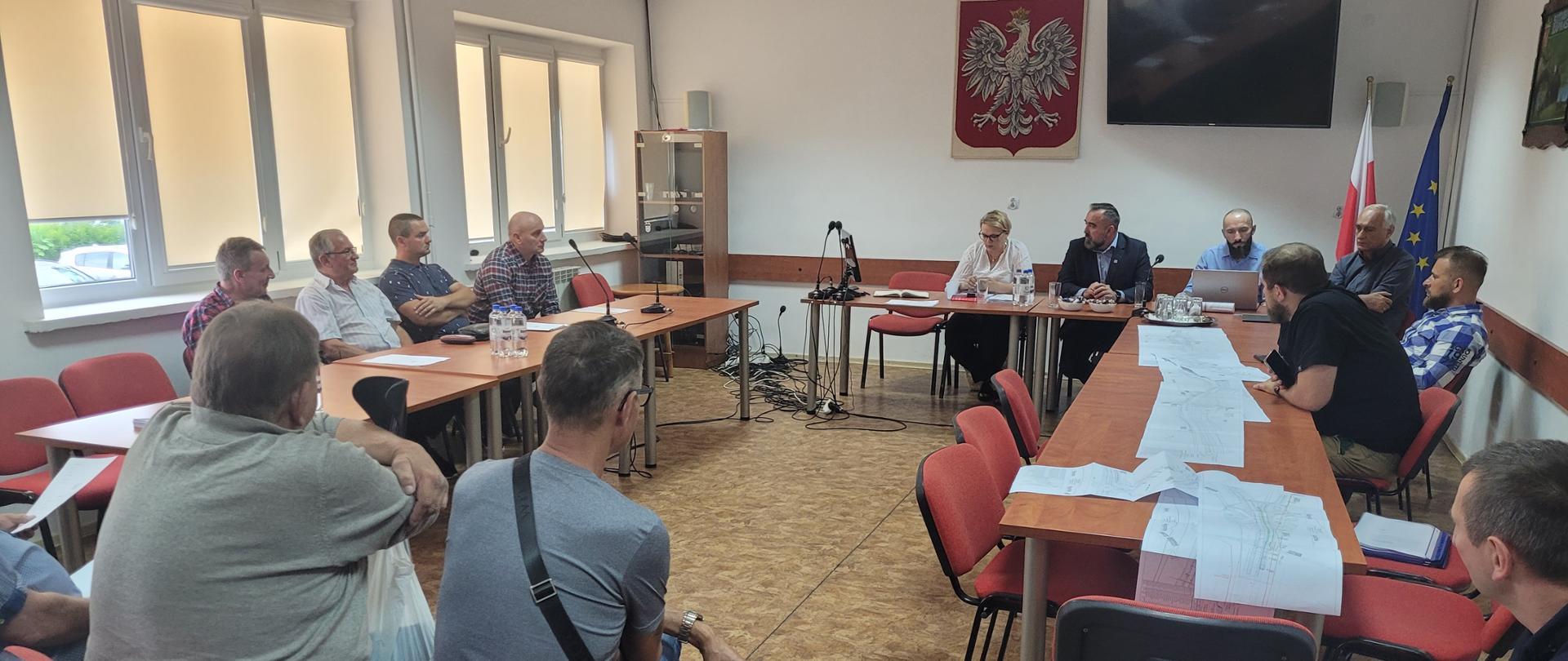 Spotkanie w pomieszczeniu, na ścianie zawieszone duże godło Polski, pod godłem, za stołem siedzi Pan Dyrektor GDDKiA Oddziału w Białymstoku i Pani Naczelnik Wydziału Nieruchomości, za stołami ustawionymi wzdłuż bocznych ścian siedzą mieszkańcy i właściciele nieruchomości przeznaczonych pod budowę drogi krajowej
