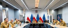 Grupa oficerów PSP siedzi po obu stronach długiego stołu konferencyjnego. Na jego końcu, przed flagami Polski i Unii Europejskiej, siedzi czterech dowódców prowadzących spotkanie.