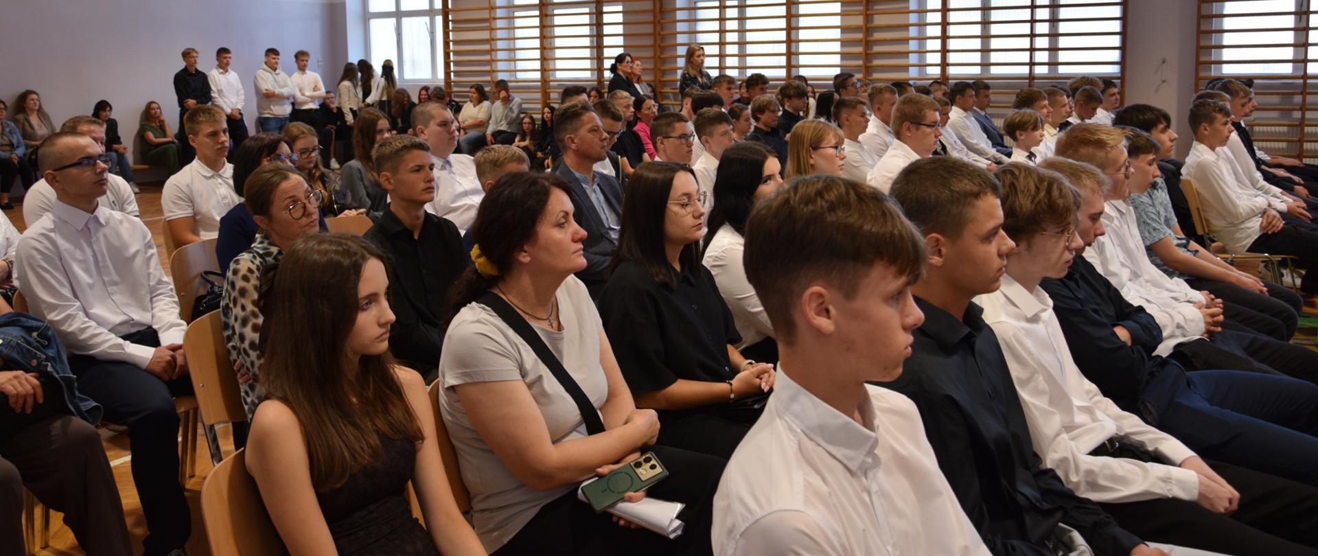 Uczniowie, nauczyciele i zaproszeni goście zgromadzeni w sali gimnastycznej podczas inauguracji roku szkolnego.