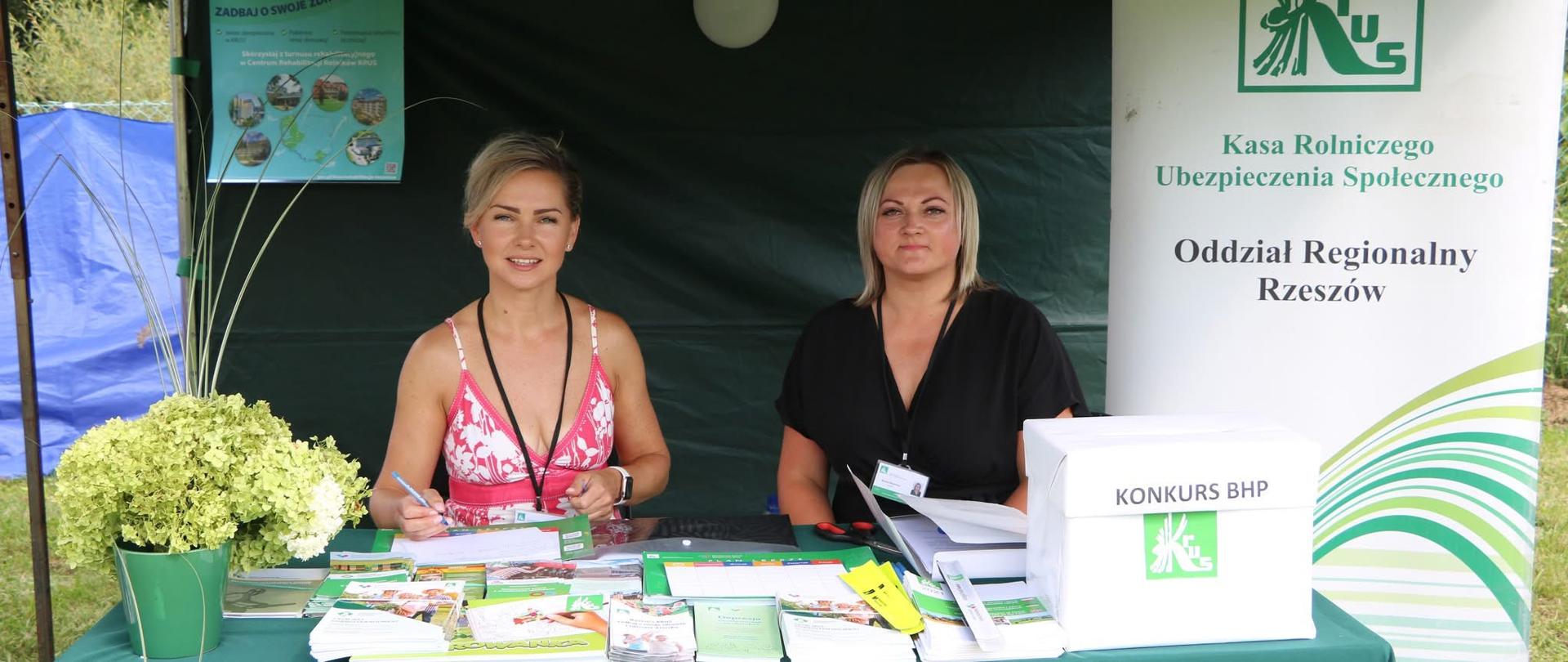 Posd zielonym namiotem, za stołem, na którym wyłożone są materiały i ulotki prewencyjne siedzą dwie kobiety. Obok stoi biały rollup z zielonym logo KRUS.