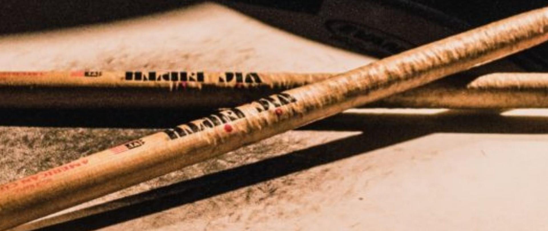 Plakat w pionie informujący o popisie uczniów klasy perkusji, który odbędzie się 10 czerwca o godzinie 18:00. Głównym elementem plakatu jest zdjęcie pałek perkusyjnych na werblu. Cały plakat w kolorystyce czerni i beżu. U dołu plakatu jasnymi listerami data i godzina wydarzenia. 