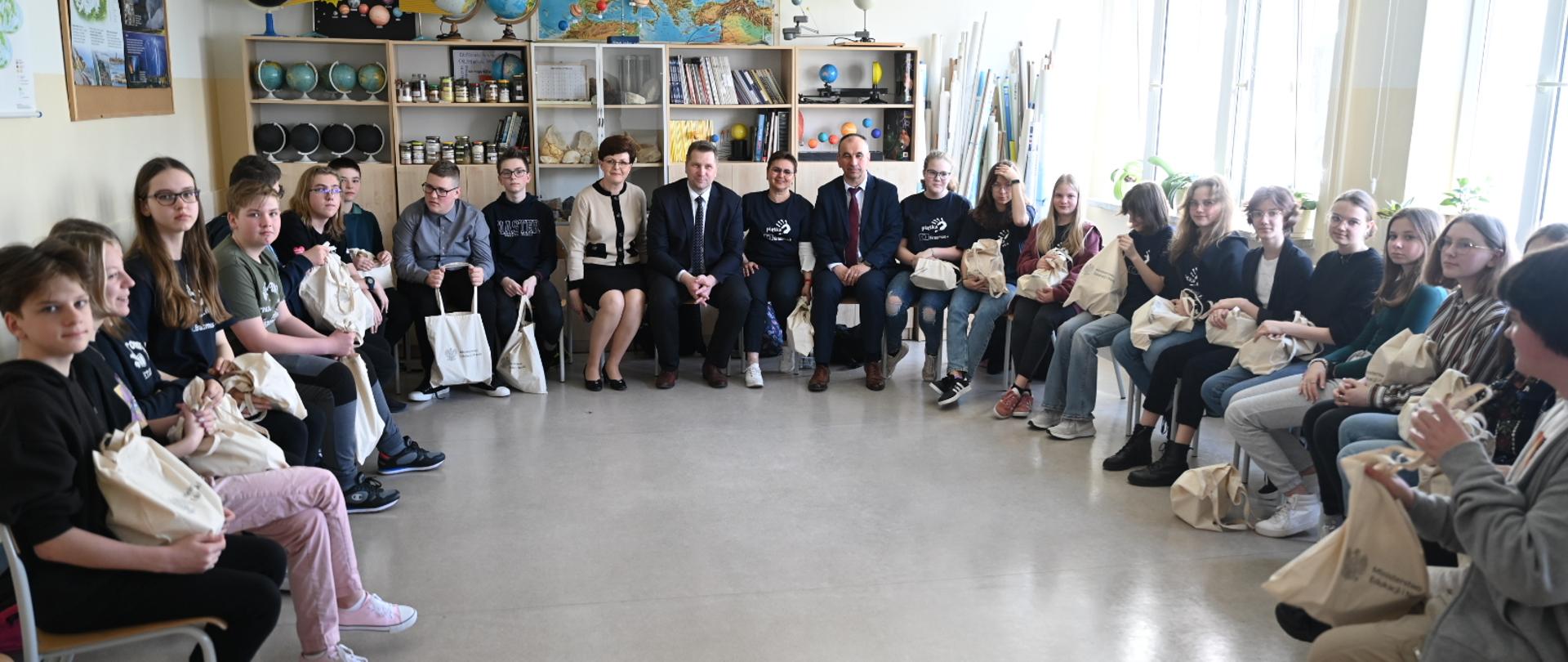 Pamiątkowe zdjęcie ministra z uczniami i nauczycielami ze świdnickiej szkoły. Wszyscy siedzą w okręgu w sali lekcyjnej.