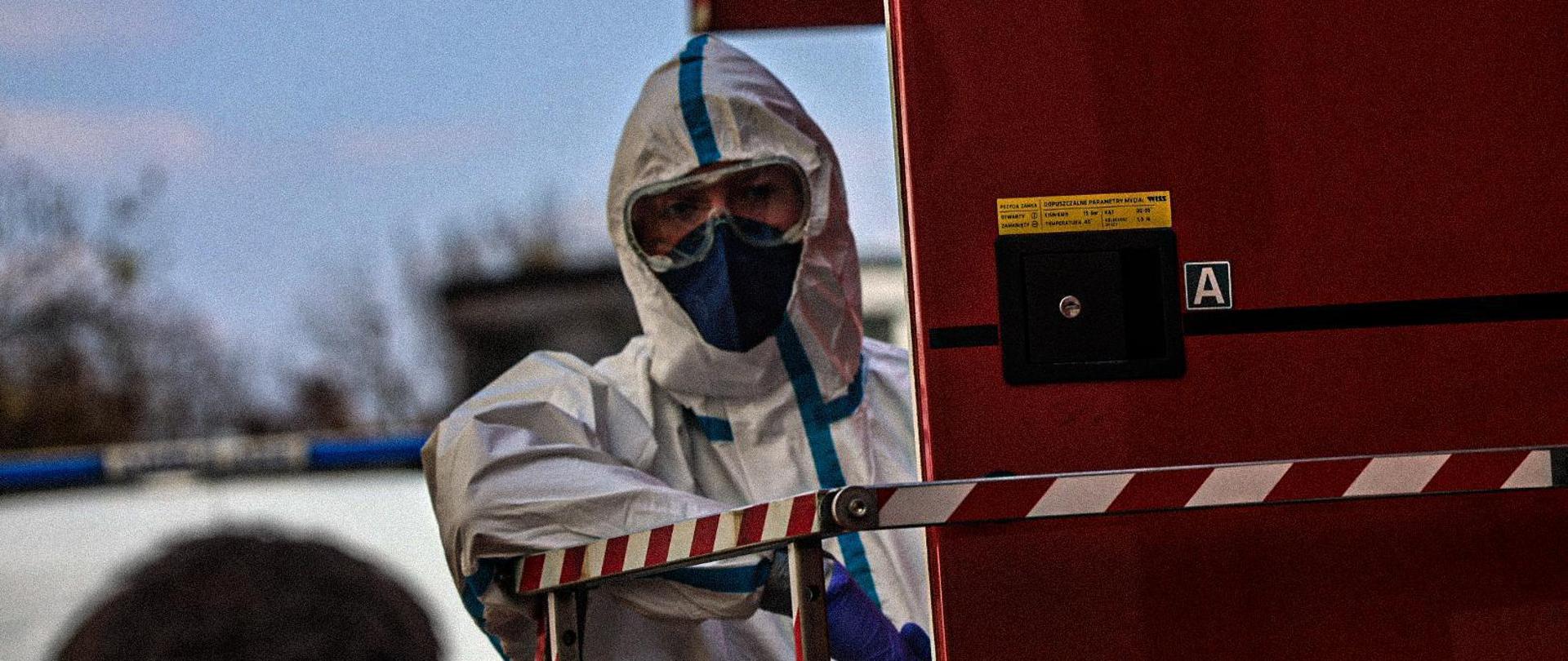 Zdjęcie przedstawia funkcjonariusza Państwowej Straży Pożarnej podczas ćwiczeń wymagających ochrony biologicznej lub chemicznej. Strażak ubrany jest w biały kombinezon z niebieskimi uszczelnieniami na szwach, ma założoną maskę filtrującą oraz gogle ochronne. Widoczne są również niebieskie rękawice ochronne.