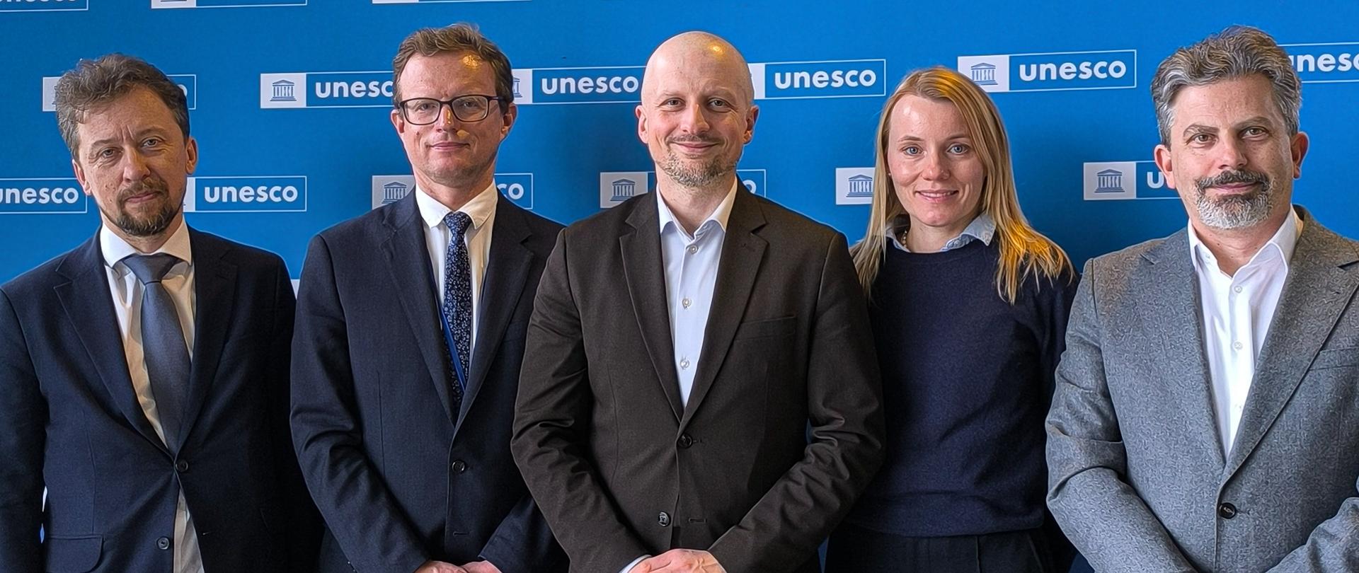 Na zdjęciu znajduje się grupa pięciu osób - czterech mężczyzn i jedna kobieta - pozujących do wspólnej fotografii na tle jaskrawoniebieskiej ścianki z wielokrotnie powtórzonym białym logo i napisem UNESCO. Wszyscy są ubrani w stroje o charakterze formalnym lub biznesowym (garnitury, marynarki, koszule). Stoją obok siebie, uśmiechając się lekko w stronę aparatu. 