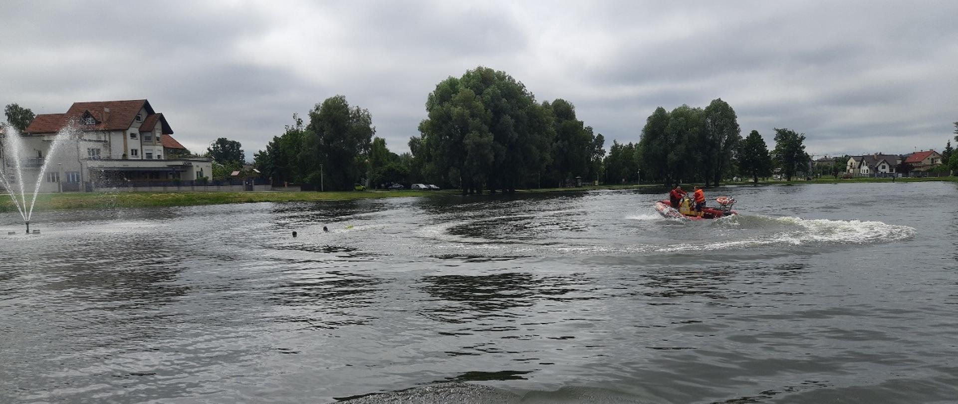 Zdjęcie obrazuje akwen łódź z ratownikami w tle drzewa i budynki